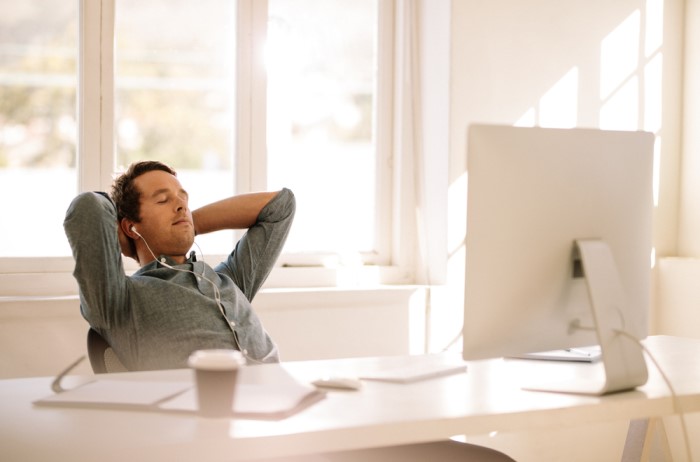 Een man zit aan een bureau met computer, terwijl hij met zijn ogen dicht achterover leunt met zijn handen achter zijn hoofd gevouwen.